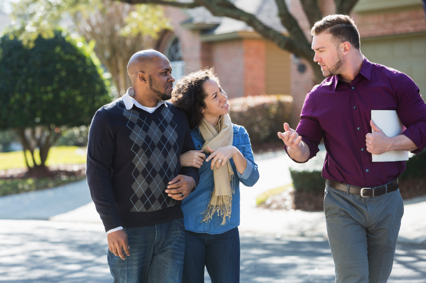 An African American couple walking on a residential street, with a young man holding a folder. He is a real estate agent, talking to them about the houses in the neighborhood.